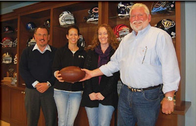 With helmets from their favorite teams – and favorite construction partners – in the background, Sports Construction Group president and CEO Paul Franks, right, hands the ball off to a trio of teammates, executive vice president George Pfeffer, vice president Julie Cipolla and controller Kelly Kost.