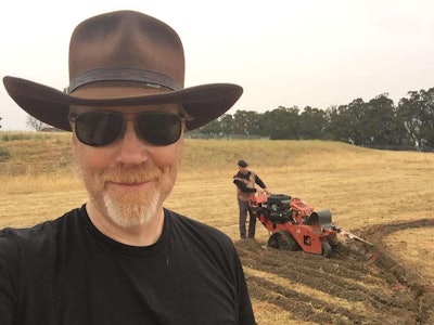 Mythbusters host Adam Savage snaps a selfie with co-host Jamie Hyneman working with a Ditch Witch trencher in the background.