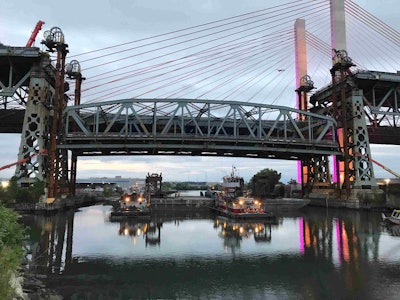 The Skanska-led joint venture team lowers the 5 million pound span of the former Kosciuszko Bridge onto two barges below, with the new Queens-bound Kosciuszko Bridge illuminated in the background. (Skanska USA)