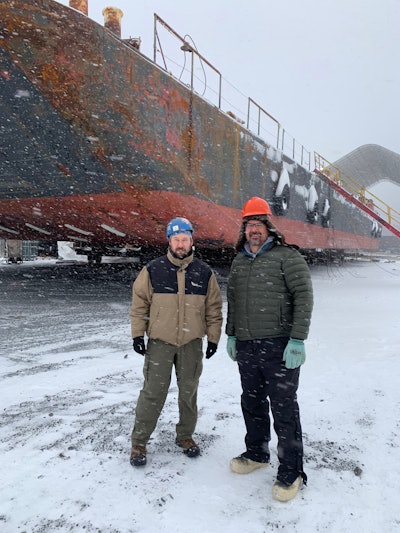 Toby Drake (right) and Daniel Paajanen, the company chief technology expert, next to one of the barges they will use to transport equipment and materials up the coast of Alaska once the sea ice thaws.