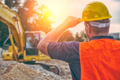 Construction worker holding hard hat looking at excavator at sunset.