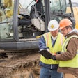 Two workers looking at tablet next to excavator
