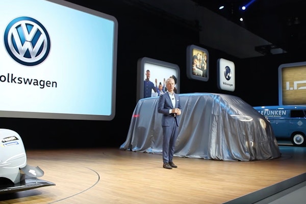Scott Keogh at the LA Auto Show in 2018 beside VW bus draped in silver cover with VW symbol on screen in background