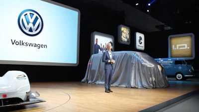 Scott Keogh at the LA Auto Show in 2018 beside VW bus draped in silver cover with VW symbol on screen in background