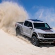 Stock 2023 Ford F-150 Raptor R pickup truck in avalanche gray shoots up sand behind it driving down dune