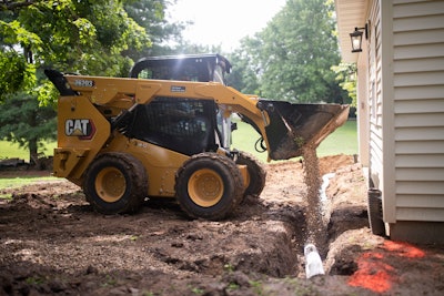 Cat 262D3 skid steer dumps gravel over pipe in trench beside house