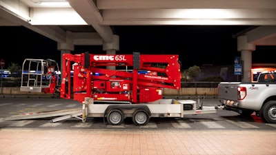 red CMC 65 L compact, tracked aerial lift in stored position on trailer behind pickup