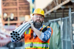 older bearded worker in yellow hard hat carrying pipes on jobsite