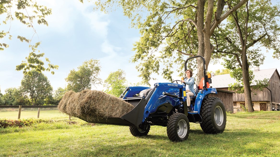 Blue farm tractor with front-end loader carrying hay bales in a rural field with trees