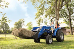 Blue farm tractor with front-end loader carrying hay bales in a rural field with trees