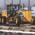 Yellow John Deere motor grader parked at industrial facility with corrugated metal building in background