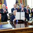 Official signing ceremony at the Resolute Desk with applauding officials and flags in background