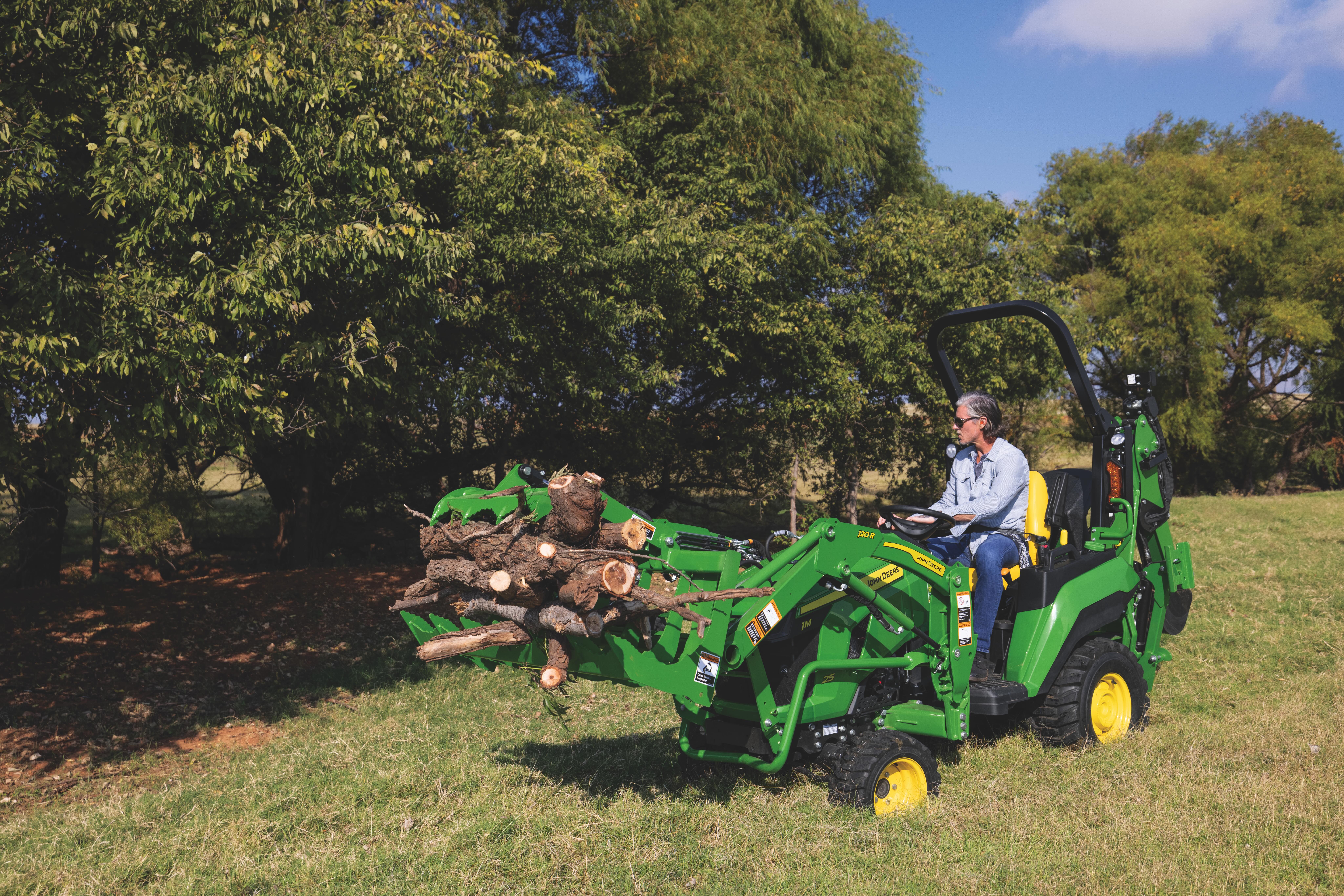 Green John Deere tractor moving logs in grassy field with trees