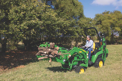 Green John Deere tractor moving logs in grassy field with trees