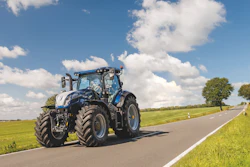 Blue agricultural tractor driving on a country road with green fields and cloudy sky