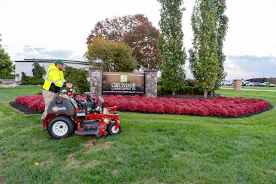 Grunder Landscaping zero-turn mower on green lawn near company sign with red flower beds