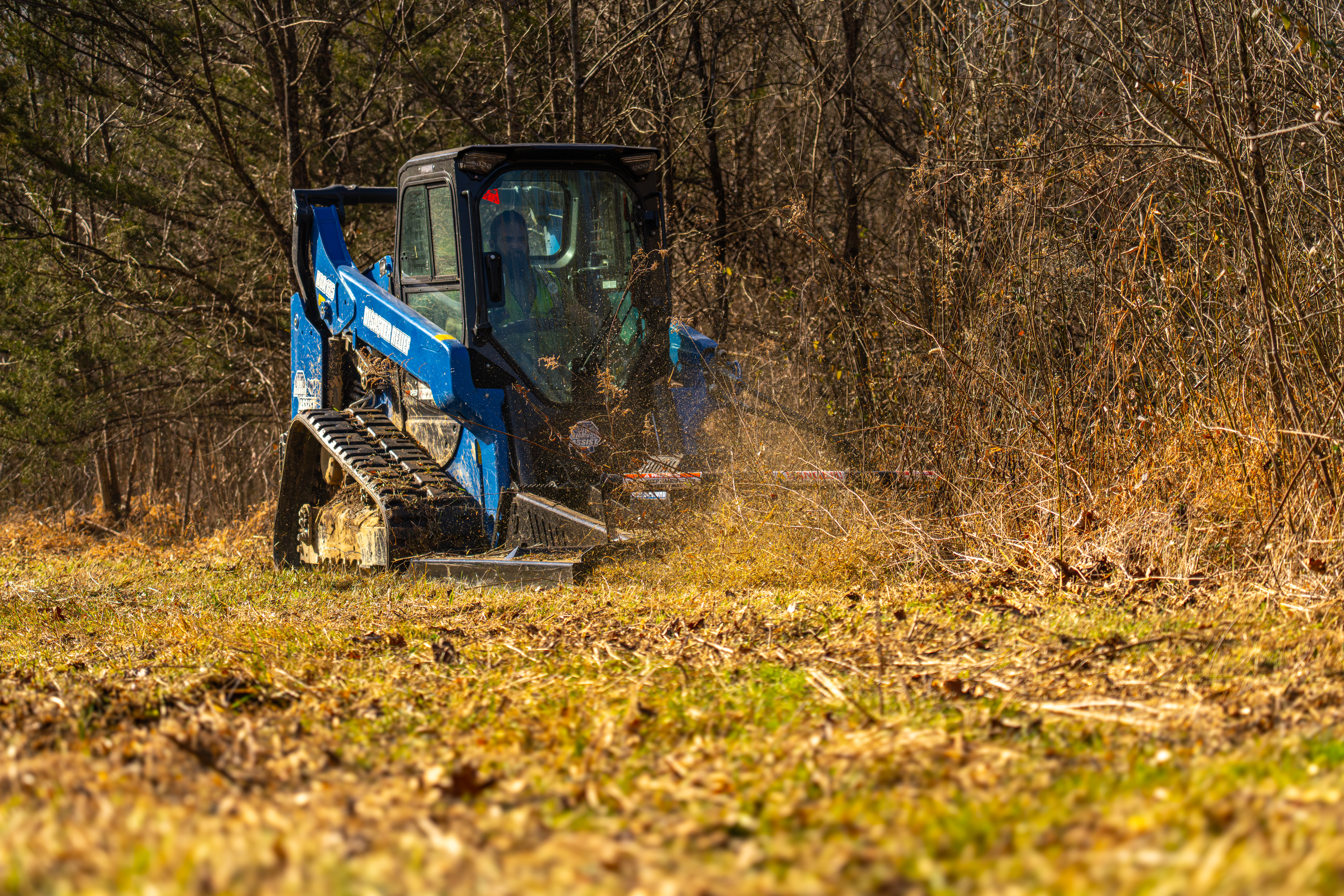 Blue compact track loader clearing brush and fallen leaves in a wooded forest area