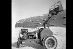 Vintage mechanical loader with articulated arm positioned in front of large corrugated metal building in snowy landscape