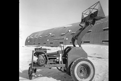 Vintage mechanical loader with articulated arm positioned in front of large corrugated metal building in snowy landscape
