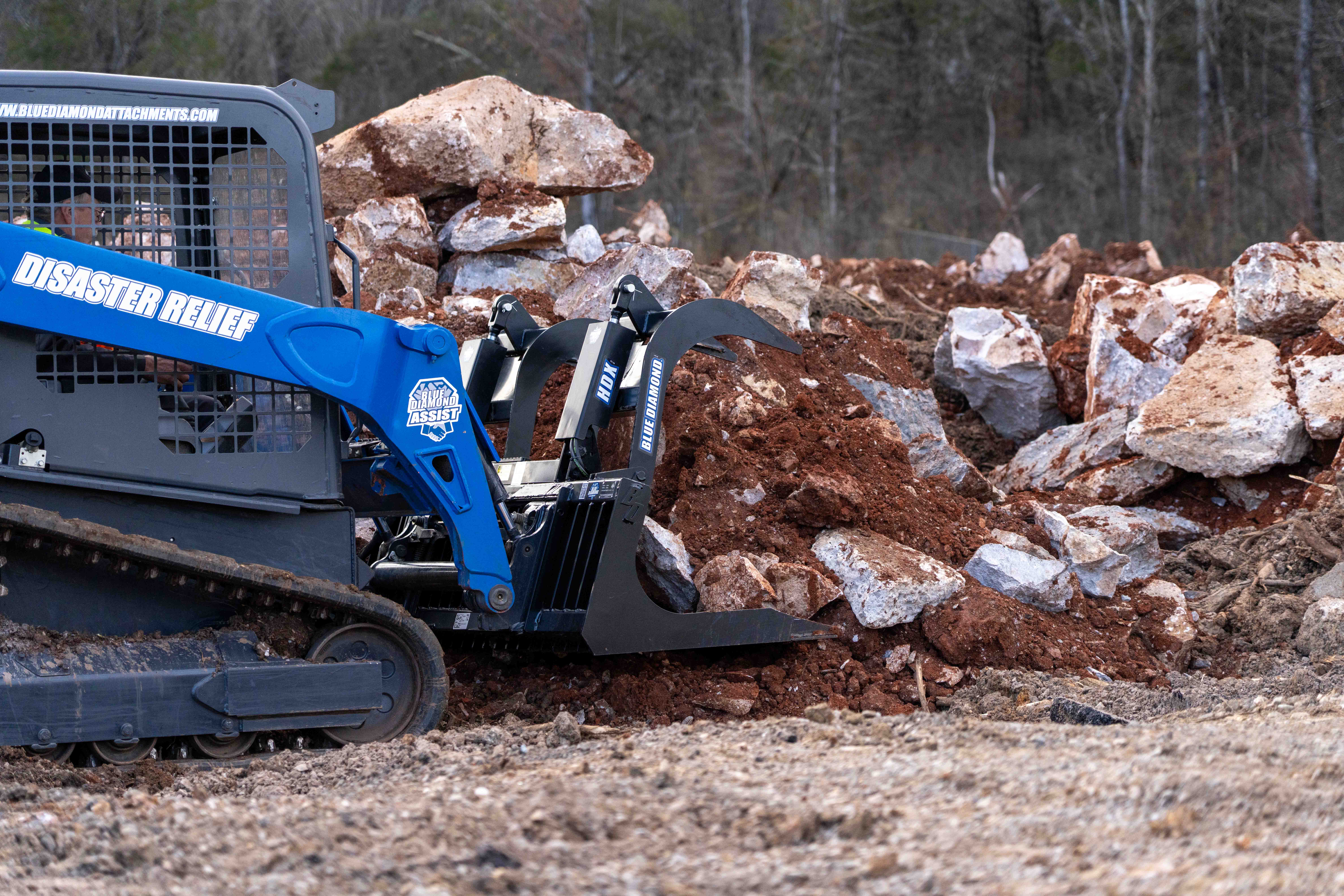 Blue skid steer loader with rock grapple attachment sorting large boulders at construction site