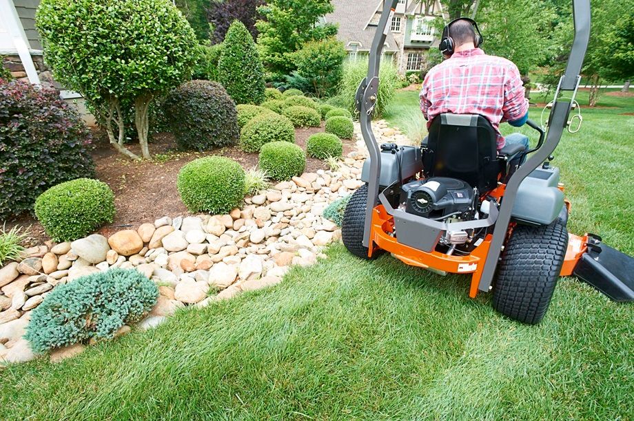 Person operating an orange zero-turn mower in a manicured residential garden with topiary shrubs and stone landscaping.