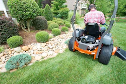 Person operating an orange zero-turn mower in a manicured residential garden with topiary shrubs and stone landscaping.