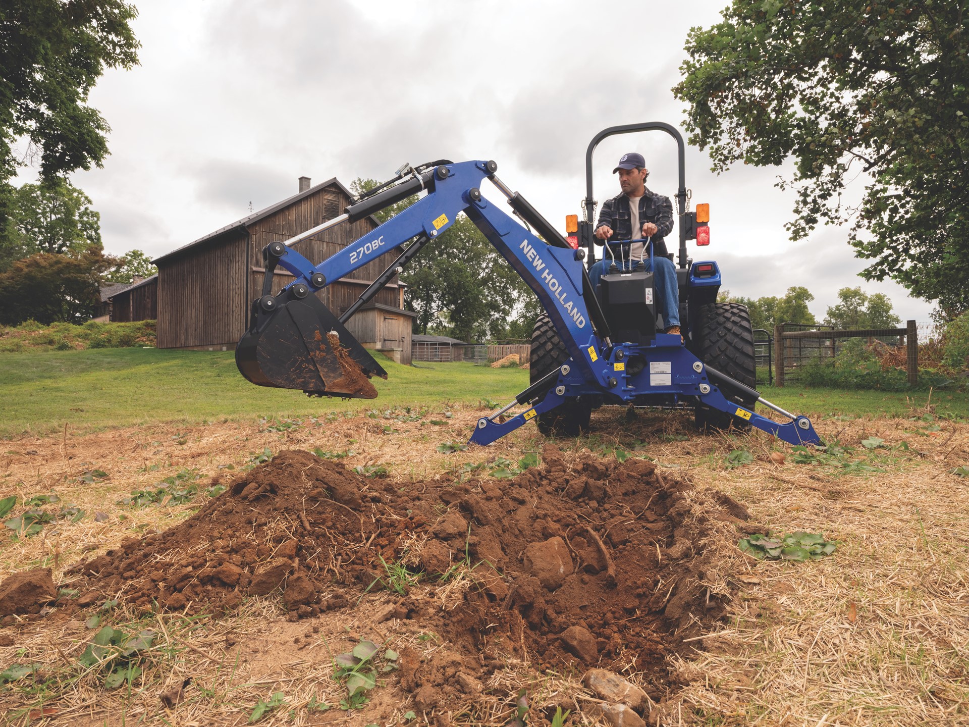 Blue New Holland mini excavator with operator digging soil on rural farm with wooden barn in background