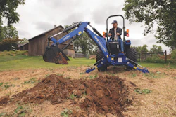Blue New Holland mini excavator with operator digging soil on rural farm with wooden barn in background
