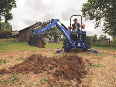 Blue New Holland mini excavator with operator digging soil on rural farm with wooden barn in background