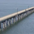 Long causeway bridge with concrete pillars extending across blue water under cloudy sky