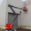 Red and black SkyJack articulating boom lift with extended arm reaching toward industrial warehouse building with metal siding