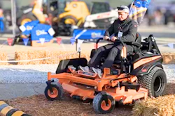 Operator demonstrates orange zero-turn radius mower at outdoor equipment show with construction machinery in background