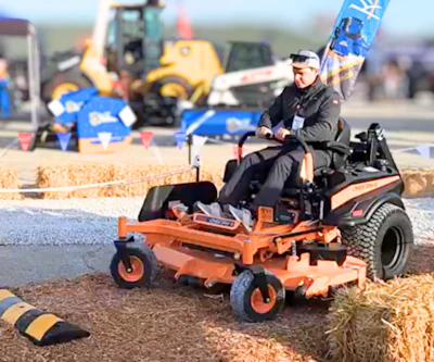 Operator demonstrates orange zero-turn radius mower at outdoor equipment show with construction machinery in background