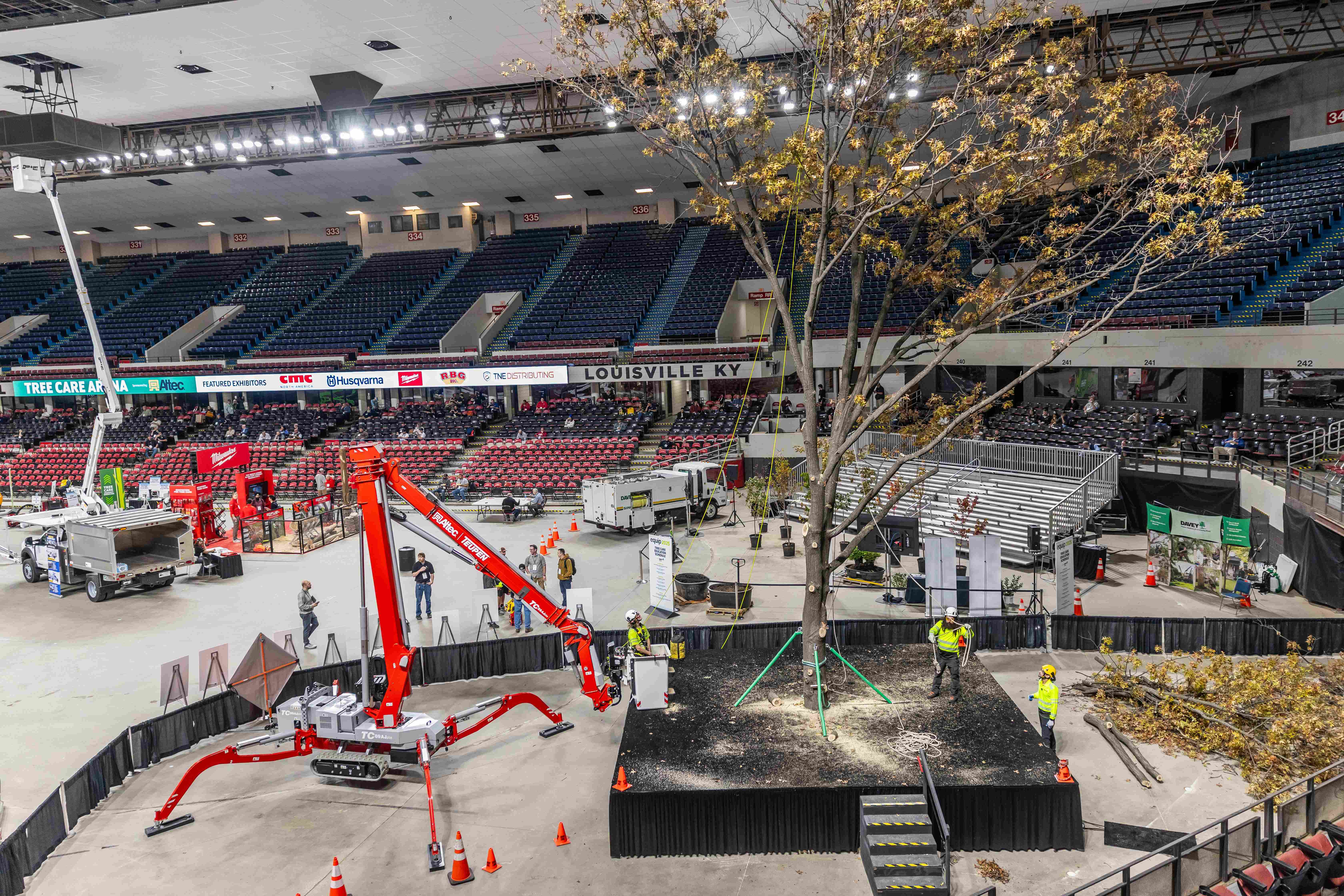 Indoor arena under construction with red cherry picker crane, temporary staging, and golden tree in foreground.