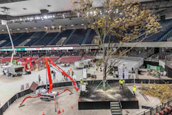Indoor arena under construction with red cherry picker crane, temporary staging, and golden tree in foreground.