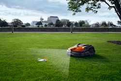 Robotic lawn mower operating on a grass field with urban buildings visible along the horizon