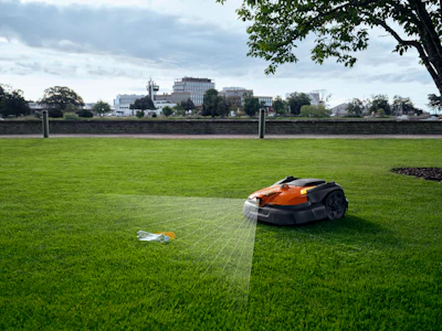 Robotic lawn mower operating on a grass field with urban buildings visible along the horizon