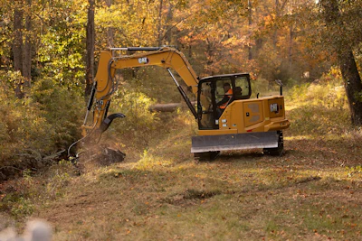 Yellow Caterpillar excavator clearing vegetation in a forested area during autumn.