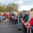 Community members and officials participate in a red ribbon-cutting ceremony at an outdoor event with autumn trees in background.