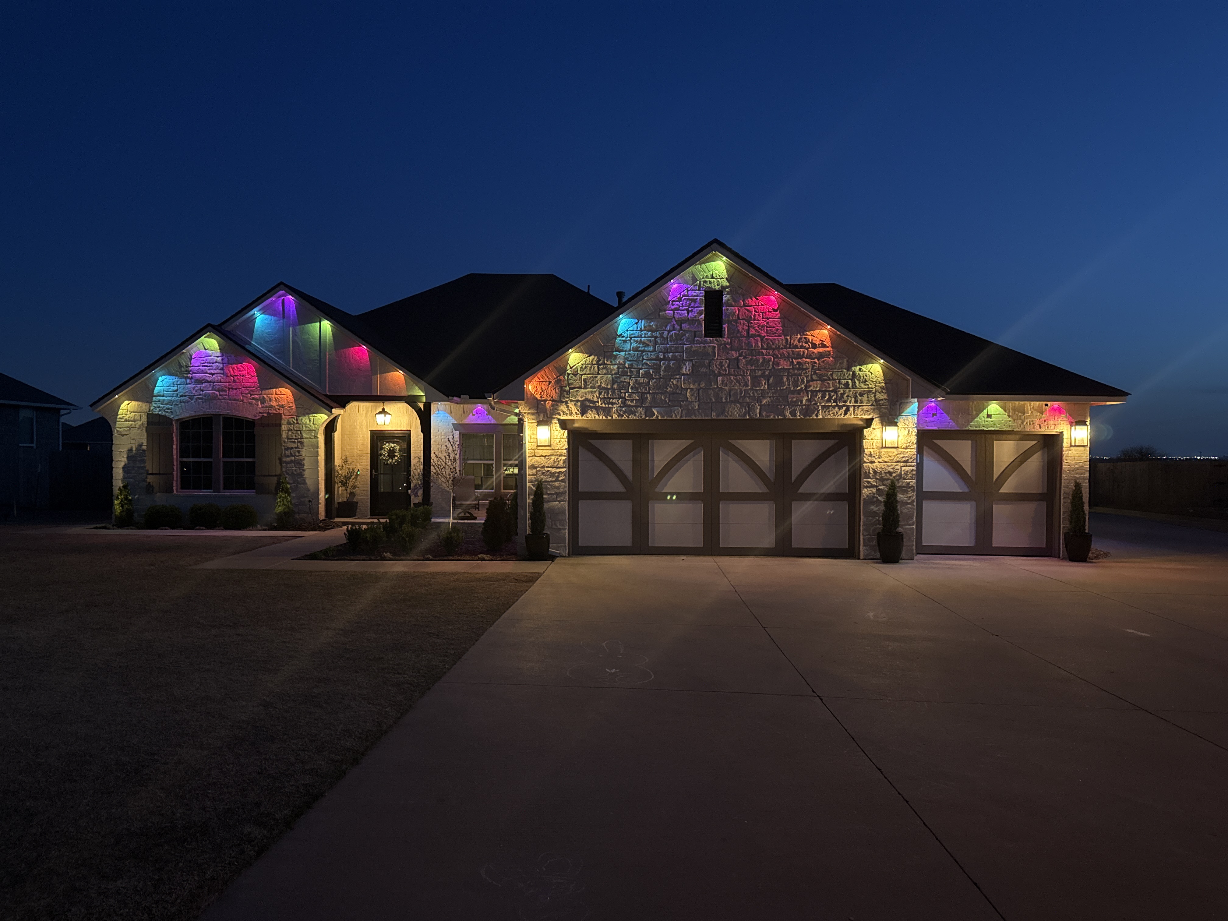 Modern stone house illuminated with colorful RGB lights at dusk, featuring three-car garage and manicured driveway