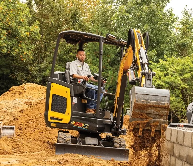 Operator using yellow and black compact excavator for earthmoving on construction site surrounded by trees