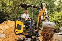 Operator using yellow and black compact excavator for earthmoving on construction site surrounded by trees