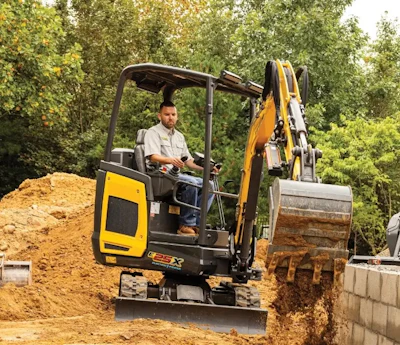 Operator using yellow and black compact excavator for earthmoving on construction site surrounded by trees