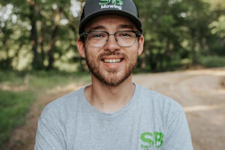 Man wearing SB Mowing branded cap and shirt smiling outdoors with trees in background