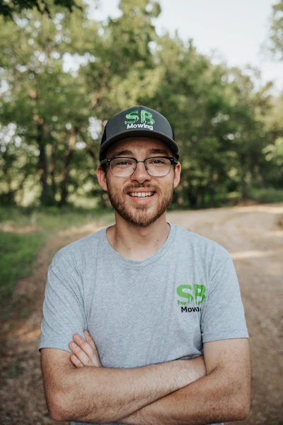 Man wearing SB Mowing branded cap and shirt smiling outdoors with trees in background