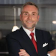 Professional headshot of a man in navy blazer and red tie smiling at camera in modern office setting