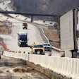 Aerial view of highway construction zone with white barriers, multiple vehicles, and winding road through wooded terrain