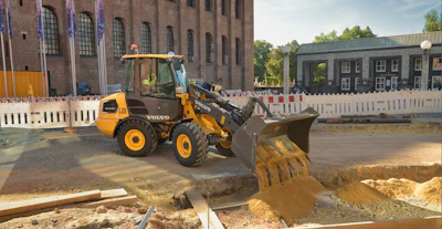 Yellow Volvo wheel loader operating at construction site with brick building and white barriers in background