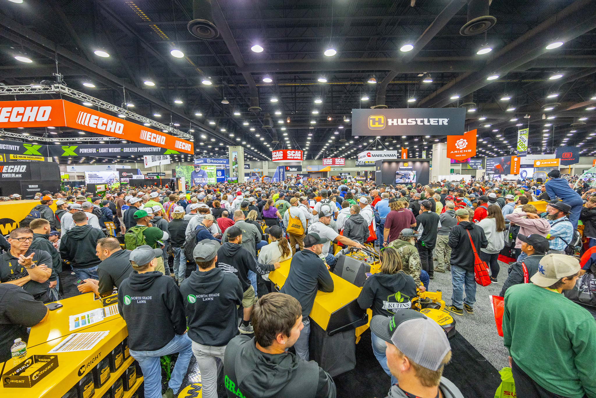 Crowded indoor trade show floor with thousands of visitors browsing equipment displays and branded booths featuring ECHO and Hustler signage.