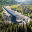 Aerial view of large construction site with blue structural framework nestled in dense coniferous forest surrounded by mountains